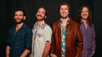 Four smiling light skinned men stand against a black backdrop.