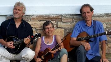 Two light skinned men and one woman sit against a stone wall. One man holds an acoustic guitar and one holds a mandolin