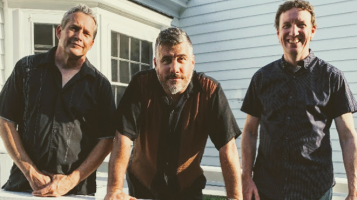 Three light skinned men stand together in front of a house with white siding
