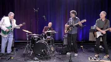 Four older light skinned men play musical instruments on a stage with a purple curtain in the background. Two play guitar, one bass, and one drums