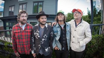 Four light skinned men stand with their arms around each other with a grey house and trees behind them.