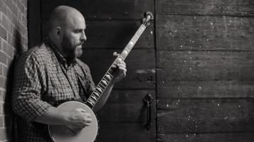 A black and white image of a light skinned man holding a banjo and leaning against a barn board wall
