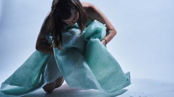 A naked light skinned woman embraces a green, crumpled glass blanket against a white background