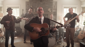 Five light skinned men play musical instruments in a living room in front of a stone fireplace. On stands front and center and wears a black suit and plays acoustic guitar. Behind him one man plays bass, two others play guitar and one plays drums