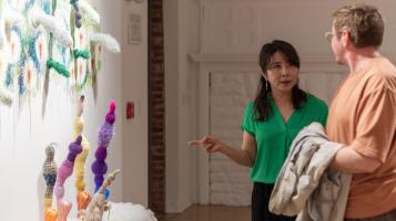 A Korean American woman with dark hair and a green blouse points to an installation of fuzzy multi colored oblong sculptural forms that spring from the wall and from a platform. She speaks to a light skinned woman with short grey hair