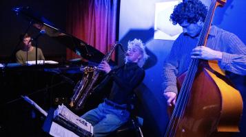 A light skinned man plays an acoustic double bass next to a light skinned woman playing a saxophone with a light skinned man playing a grand piano in the background. They are bathed in purple light