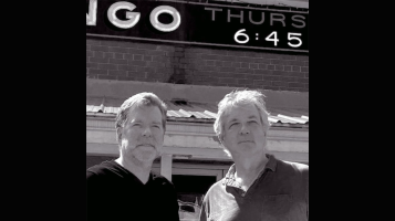 A black and white image of two light skinned men seen from the chast up standing in front of an old neon sign