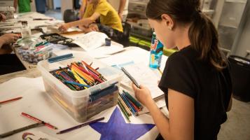 A light skinned girl seen from behind draws using a colored pencil. A bin full of colored pencils sit next to her on a table piled with papers and markers and a drawing of a large purple star. Other children are visible in the background