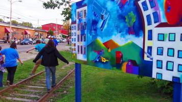 A photograph of colorful mural in the foreground depicting white buildings, green mountains and a blue sky. People walk by on old railroad tracks, grown over with grass