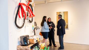 A photograph of two light skinned women and one man standing together, talking in an art gallery. A painting hangs on a white wall behind them and a soft sculpture of a red bicycle and other household items hangs on a wall in the foreground