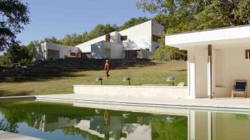 An old photo of a modern angular white house, built into a gently sloping hillside. A light skinned woman walks towards a pool and modern white cabana in the foreground.