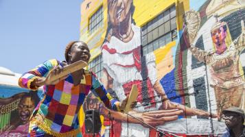 A photograph of a dark skinned woman wearing a multi colored blouse, holding two wide sticks as she smiles and dances in front of a large colorful, mural on a brick building