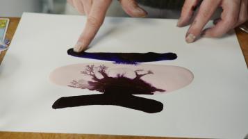 A close up photograph of an ink drawing on paper with a light skinned person's fingers pointing to a large dark blotch