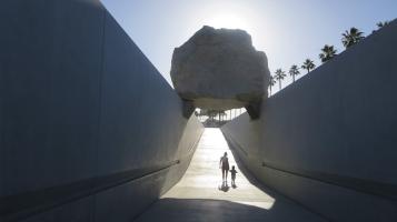 A photograph of an adult and a small child walking beneath a large craggy boulder suspended between two high walls. Palm trees are visible behind one wall with a clear blue sky in the background