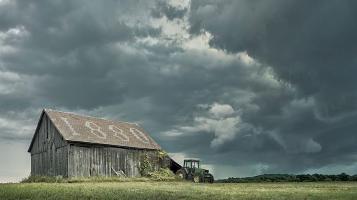 A photograph of a weathered grey barn with a roof that spells 1880 in the shingles. A green tractor is parked outside and dramatic grey clouds gather overhead.