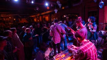 a photograph of a band playing on a low stage bathed in purple light in a bar filled with people
