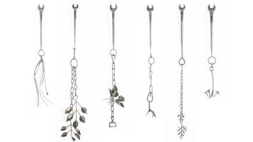 A photograph of six silver steel sculptures arranged vertically. Each work starts with a wrench that ends in a ring with danging chains that turn into flowers, vines, and other naturalistic abstract shapes