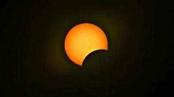 A photograph of a solar eclipse in process with an orange yellow sun, begginging to be blocked by a black sphere against a black background