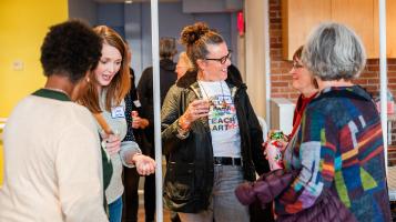A photo of five women standing together and talking in the BCA Center Gallery Learning Studio Space. One woman has dark skin and short hair and stands with her back to the camera. One has light skin and reddish brown straight hair and exclaims while holding a plastic cup. In the center of the group a woman with greyish brwn hair in a bun and light skin wears a t-shirt that says &quot;Keep Calm and Teach Art&quot; while holding a plastic cup and laughing