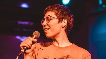 A person with short hair and glasses singing into a microphone.