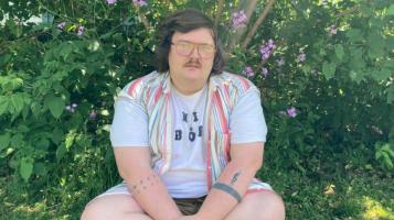 A person with glasses and a mustache and wearing a vest sits against a backdrop of green grass and bushes