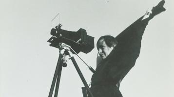 A black and white image of a woman standing behind a camera on a tripod.