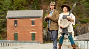 two people with big hats on, standing on a bridge with a brick building behind them.