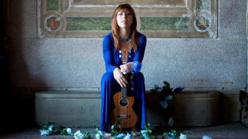 The performer sitting with her ukulele in a victorion room surrounded by blue flowers