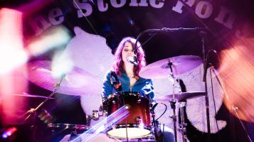 the performer sitting behind a drumkit singing into a microphone. Photo has some beautiful layered lighting effects