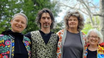 A photo of three light skinned women and one light skinned man seen from the waist up with their arms around each other's shoulders. The women wear brightly colored patterned jackets and the man wears a patterned vest.