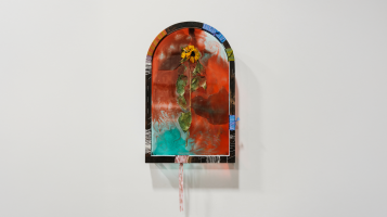 A photograph of a wall hanging sculpture by artist Lydia Kern, called Double Sorrow, Double Joy. The sculpture is shaped like an arched window and is made of colorful acrylic and flowers