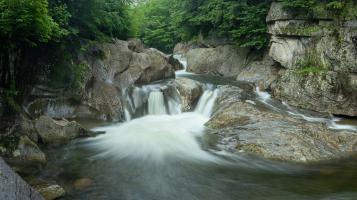 Photograph of a short rocky waterfall in primarily greys and vibrant greens for the forest scenery behind and around. 