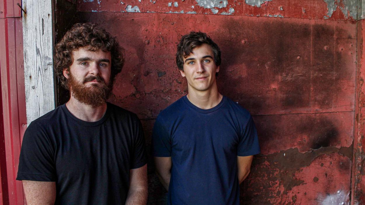 2 Band members in t-shirts (one with a scruffy beard) standing in front of a distressed wall