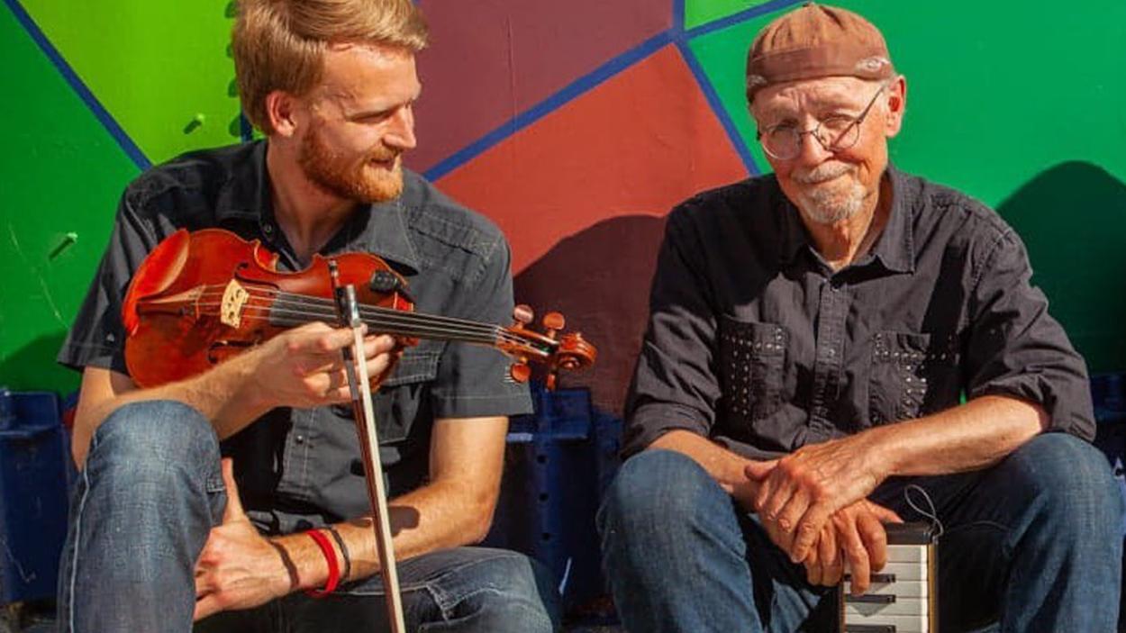 Pete and Oliver holding a violin and keyboard in front of multi-color geometric wall