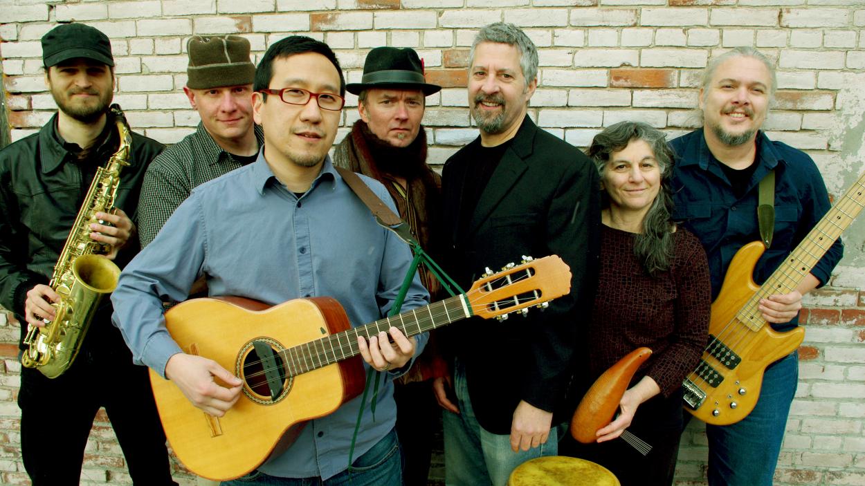 The members of GuaGua stand in front of a brick wall holding musical instruments.