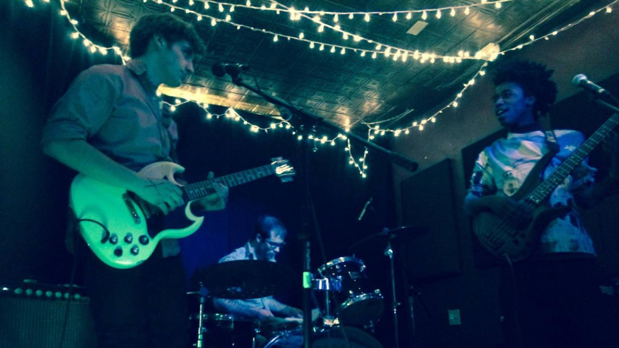 The three members of Binger on a stage strung with white twinkle lights, one holds a guitar, one holds a base, one sits behind drum set.