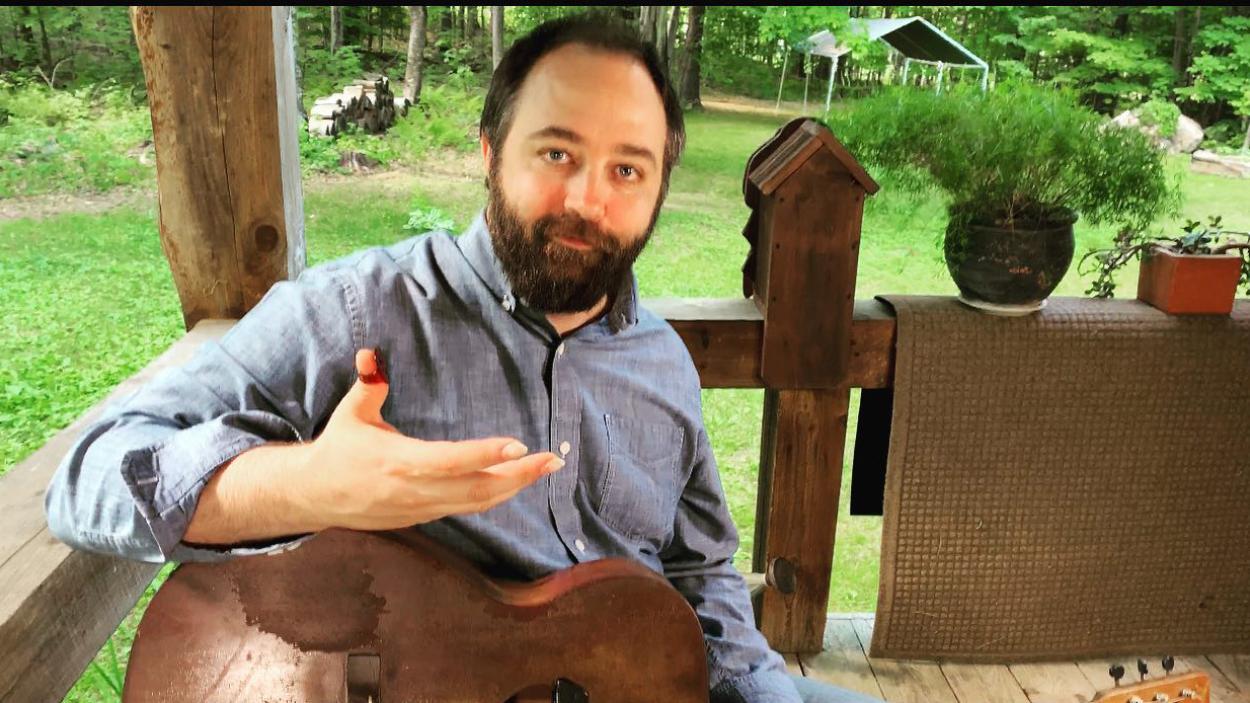 Zak Trojano holds a guitar as he sits on a porch, with green grass in the background.