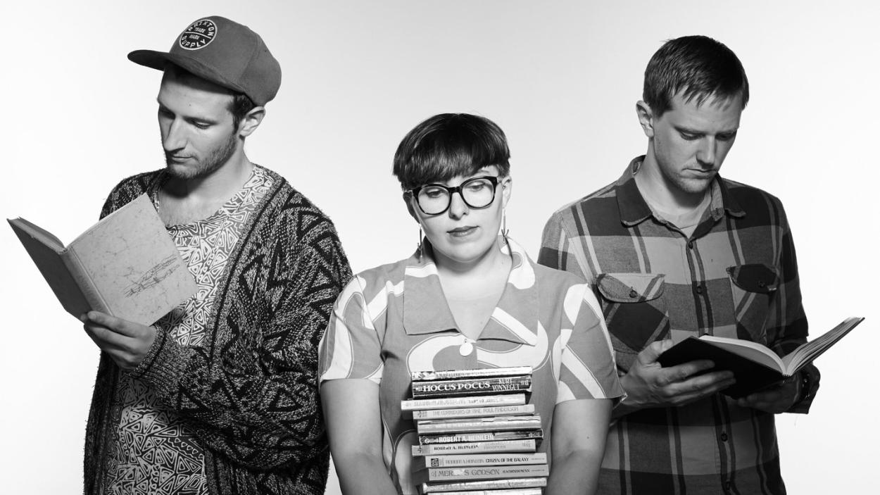 The three members of the Leatherbound Books, seen in black and white, stand together, as two of them read books and one holds a large stack of books.
