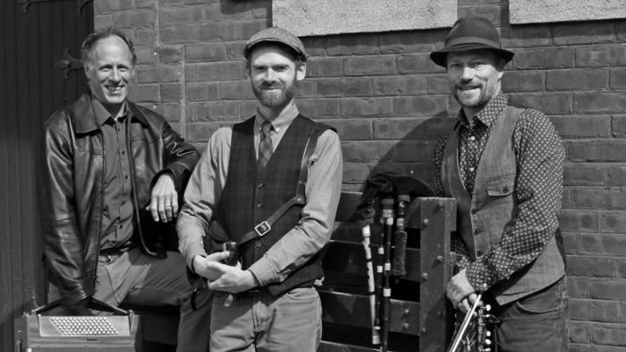 The three members of Triton stand in front of a brick wall, one holds a fiddle, and one holds an accordian.