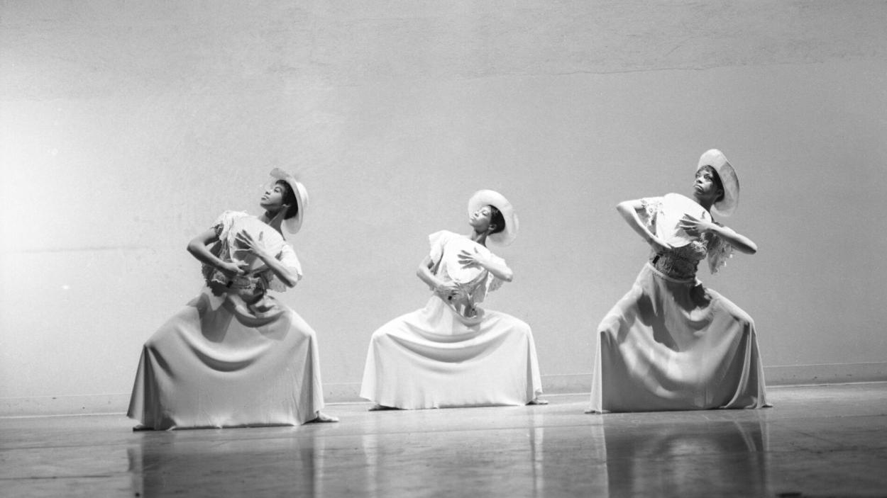 Three dancers from the Alvin Ailey company plié while holding fans and wearing long dresses and sunhats.