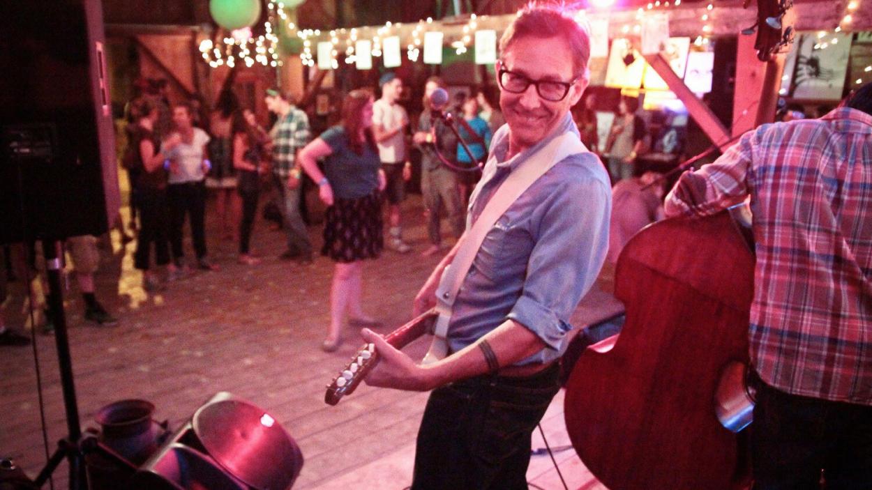 Brett Hughes wears a light blue shirt and holds an electric guitar, with an audience on onlookers in the background.