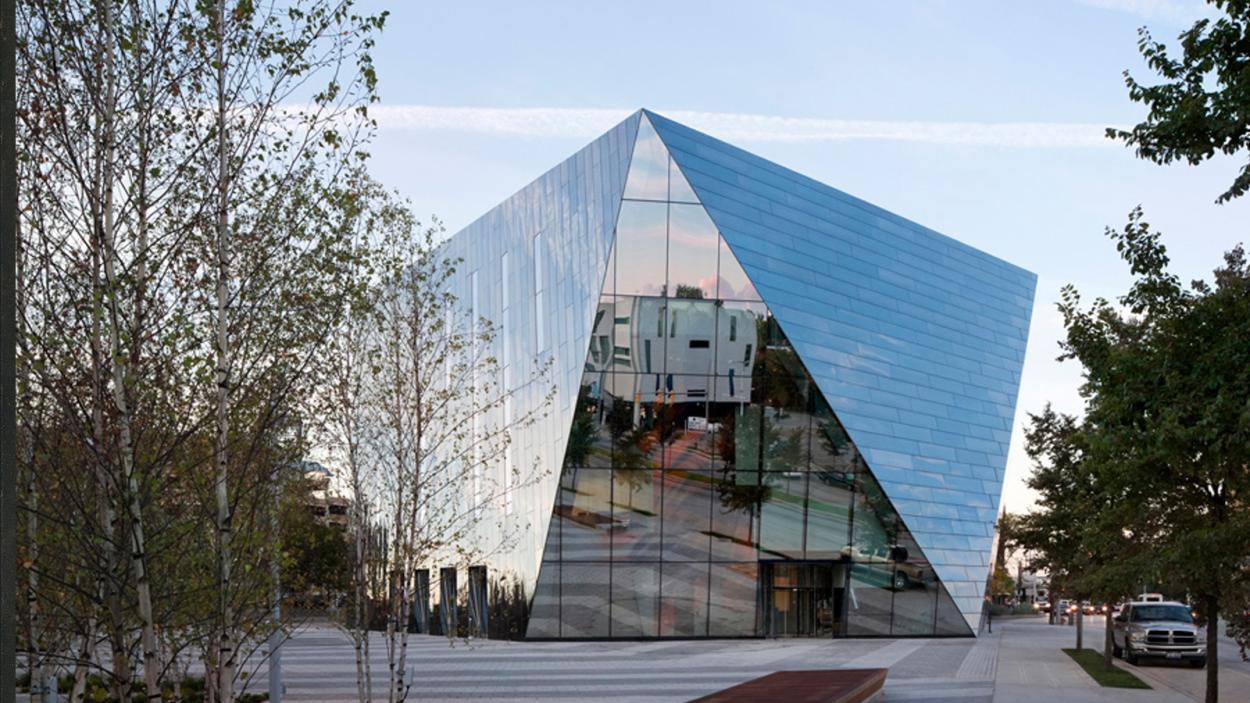 An angular building covered with reflective glass windows, with birch trees and a city street in the foreground.