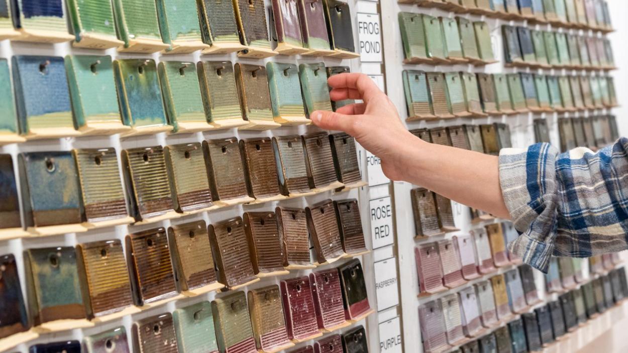The hand of a light skinned person wearing a plaid shirt reaches toward a wall of clay tiles with various colors of glazes