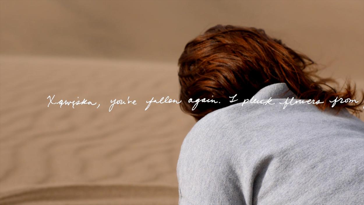 A still from a video showing a women's head and shoulders, leaning forward, as seen from behind with white cursive script overlaid that says Xąwįska you're fallen again. I pluck flowers from