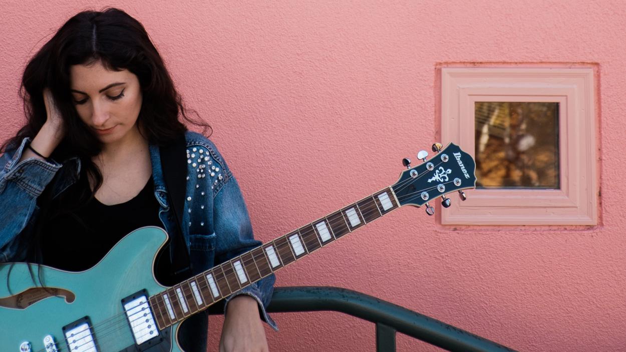 Ali T holding a vintage teal guitar in front of a pink concrete wall