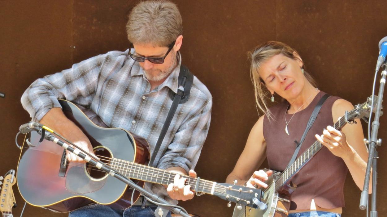 Dana is playing acoustic guitar and Beth is playing a banjo onstage, both have their eyes closed