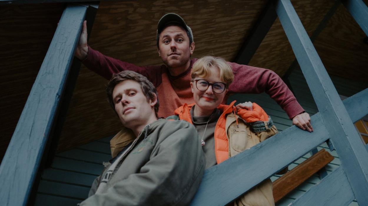 Two light skinned men and one light skinned woman with short blonde hair and glasses stand close together on an outdoor staircase with blue handrails, as seen from below.