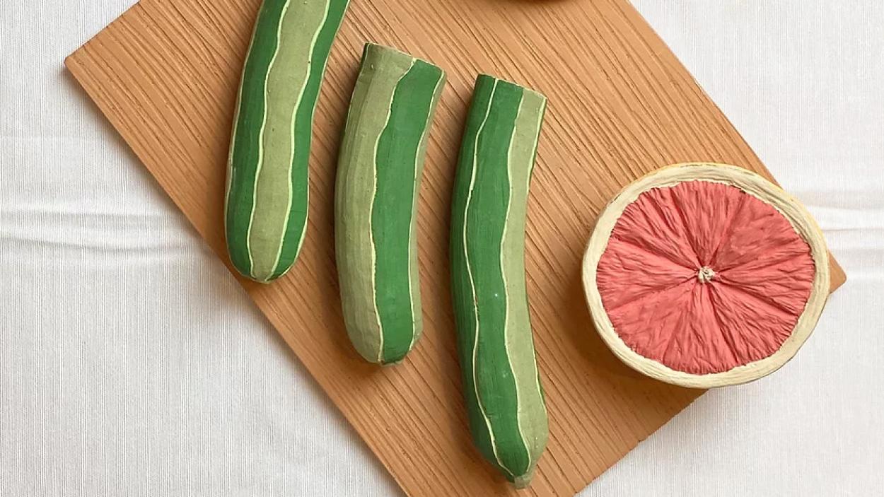 A photograph of a ceramic sculpture of half of a sliced grapefruit and three sliced cucumbers arranges on a cutting board