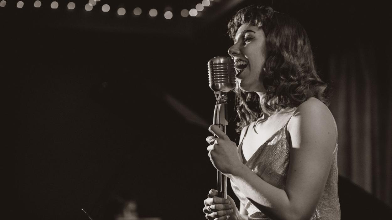 A black and white photograph of a light skinned woman with shoulder length dark, wavy hair singing into a microphone