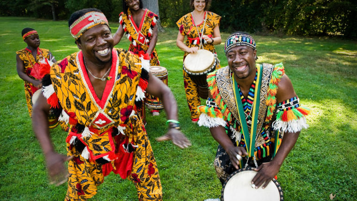 In the foreground, two dark skinned men stand on a green lawn, wearing colorful traditional African clothes, while one play a drum and one dances. In the background two dark skinned women and one light skinned women wear colorful traditional African clothing and play drums
